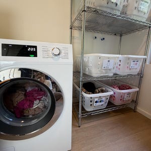 Op de afbeelding: Een witte wasmachine met voorlader en een digitaal display dat 2:35 aangeeft, naast een hoog metalen draadrek. Het rek bevat verschillende witte plastic wasmanden, elk voorzien van een label met tekst voor het sorteren van de was.