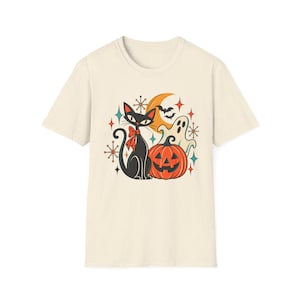 May include: A cream-colored short-sleeve t-shirt featuring a graphic print of a black cat with a red polka-dot bow, an orange jack-o'-lantern, a white ghost, a crescent moon, and small starburst shapes in shades of orange, teal, and black.
