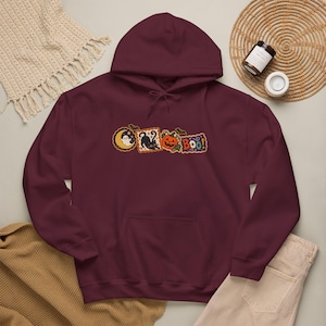 Peut inclure: Un sweat à capuche bordeaux avec une impression graphique centrale représentant un fantôme sur une lune jaune, un chat noir, une citrouille d'Halloween orange et le texte "BOO!" en orange. Le sweat possède des manches longues et une poche kangourou.