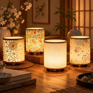Può includere: Quattro lampade da tavolo cilindriche con paralumi in tessuto poggiano su una superficie in legno. Un paralume presenta un motivo a fiori bianchi e oro, uno una stampa floreale colorata, uno è bianco sporco e uno mostra un delicato disegno floreale rosa e blu.
