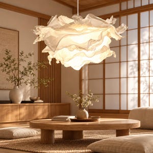 Peut inclure: Une suspension lumineuse blanche composée de multiples couches de tissu translucide et volanté créant une lueur douce et diffuse. L'abat-jour présente un aspect volumineux semblable à un nuage avec des bords organiques irréguliers.