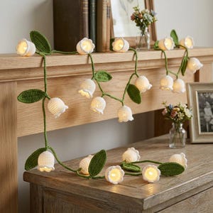 May include: A string of decorative lights featuring crocheted white lily of the valley flowers and green leaves. The flowers are shaped like small bells, each containing a warm glowing light bulb, connected by a green crocheted vine.