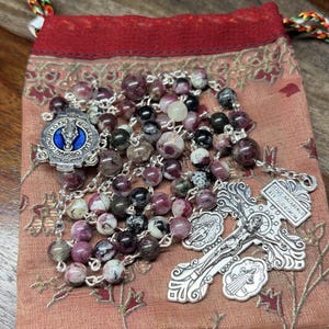 May include: A rosary featuring multi-colored purple, black, and white stone beads on a silver-tone chain. It includes a silver-tone crucifix with the text Jesus Nazarenus Rex Judaeorum and a center medal with a blue enamel inlay.