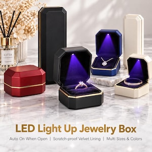 May include: A collection of octagonal jewelry boxes in black, red, blue, and cream, each featuring an interior LED light that illuminates when opened. The boxes have gold-tone trim and dark velvet linings, displaying rings and a necklace.