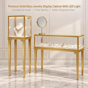 May include: A gold-framed glass jewelry display cabinet with LED lighting, featuring a tall pedestal unit with a necklace bust and a long horizontal counter unit with a round mirror and various rings and watches on display inside.
