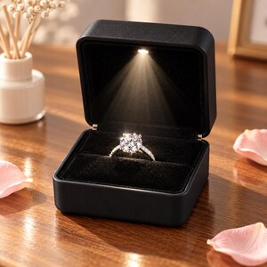May include: A square black ring box with a velvet interior and a built-in LED light that illuminates a silver-toned ring featuring a large round-cut clear stone and smaller accent stones along the band, centered within the open display case.
