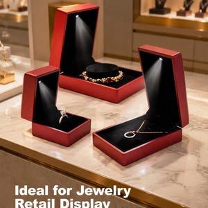 May include: Three red jewelry boxes with black velvet interiors and integrated LED spotlights. One box holds a diamond ring, another a multi-colored gemstone bracelet, and the third a circular pendant necklace. The text reads: Ideal for Jewelry Retail Display.