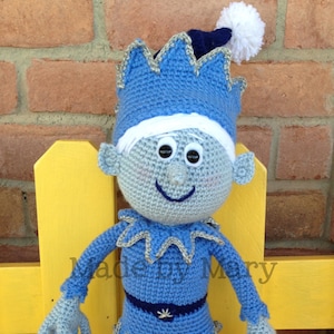 Peut inclure: Poupée au crochet faite à la main en forme d'elfe d'hiver. La poupée est principalement bleu clair avec des accents bleu foncé, un chapeau à pompon blanc et un flocon de neige. Le texte "Made by Mary" est visible.