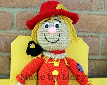 May include: A crocheted scarecrow doll with a red hat, yellow hair, and blue pants. The scarecrow is holding two black birds. The doll is sitting on a yellow bench. "Made by Mary" is written on the doll's shirt.
