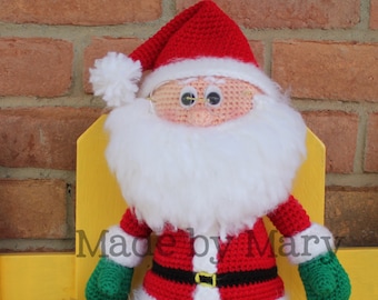 Pode incluir: Um boneco de Papai Noel de crochê vestindo um terno vermelho, detalhes brancos e uma barba branca. Ele está segurando um saco verde com um laço amarelo. O boneco está sentado em um banco amarelo. Feito por Mary.