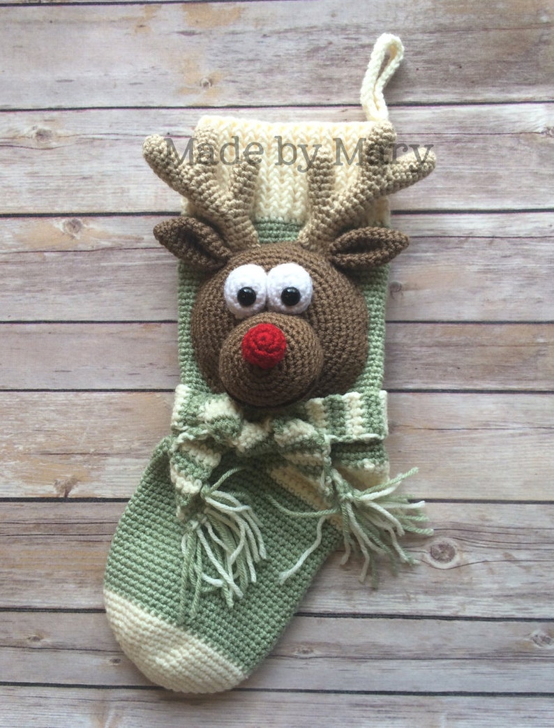 May include: A green and white crocheted Christmas stocking with a reindeer face. The reindeer has brown fur, big eyes, a red nose, and antlers. The stocking has a white and green striped scarf with tassels.