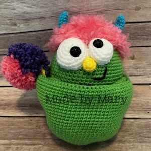 May include: A green crocheted monster with pink and purple pom-poms, white eyes, a yellow nose, and a blue and pink fuzzy head. The monster is sitting on a wooden surface and has the text "Made by Mary" on its body.