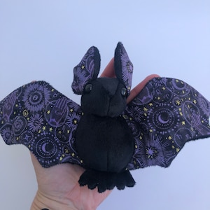 May include: A black plush bat with purple and gold wings. The wings are patterned with stars, moons, and other celestial symbols.