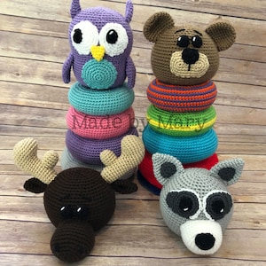Puede incluir: Un conjunto de cuatro juguetes de animales de crochet, que incluyen un búho, un oso, un alce y un mapache. El búho y el oso están encima de anillos de apilamiento coloridos.