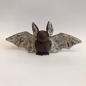 May include: A brown plush bat with beige wings. The wings are printed with black illustrations of various baking ingredients and tools, including a whisk, a rolling pin, a knife, and a loaf of bread.