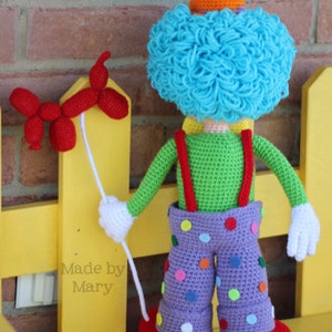May include: A crocheted clown doll with a blue curly wig, orange hat, green shirt, purple pants with colorful polka dots, and red shoes. The clown is holding a red balloon animal.