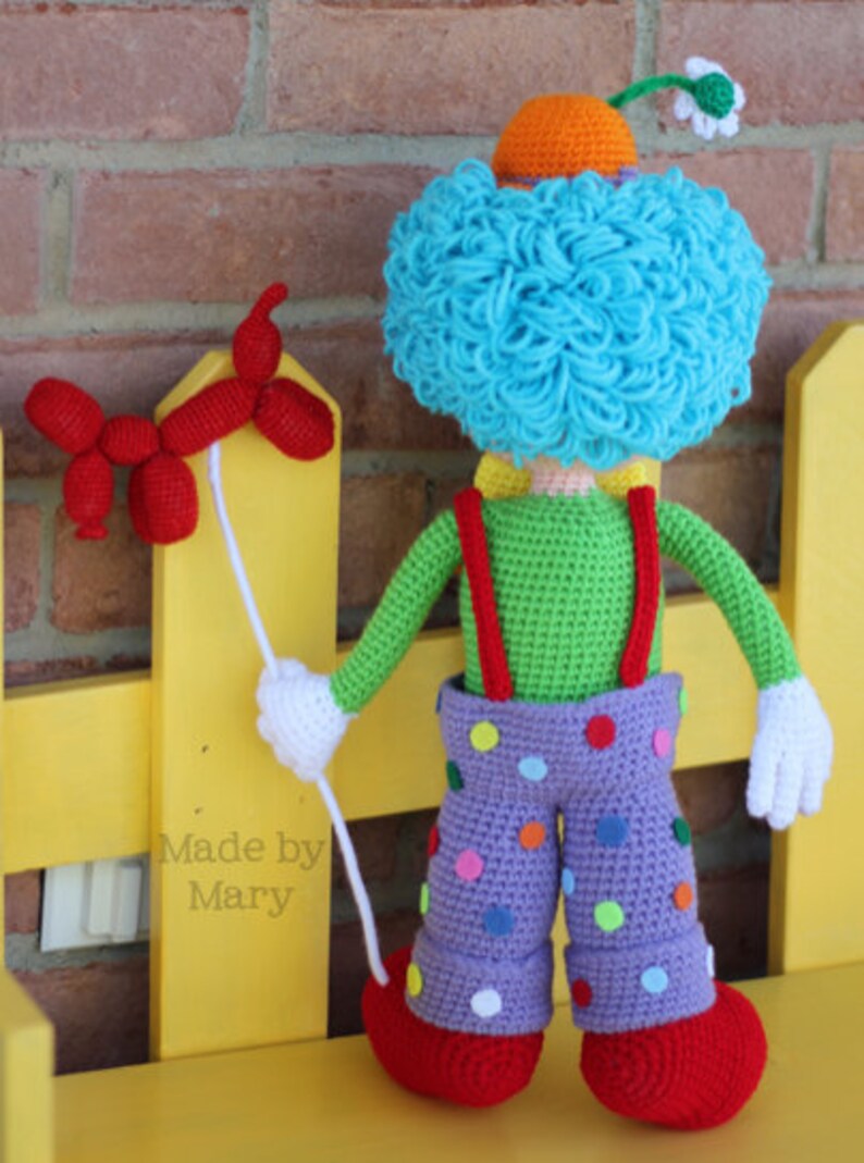 May include: A crocheted clown doll with a blue curly wig, orange hat, green shirt, purple pants with colorful polka dots, and red shoes. The clown is holding a red balloon animal.