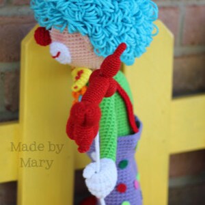 May include: A crocheted clown doll with blue hair, a red nose, and a colorful outfit. The doll is holding a white string and is standing on a yellow surface. The text "Made by Mary" is visible in the image.