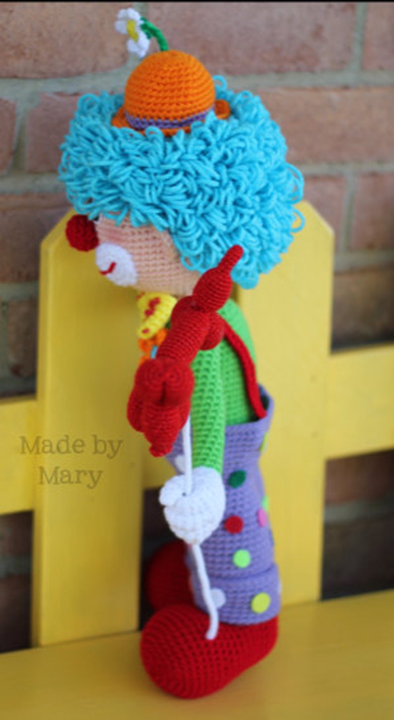 May include: A crocheted clown doll with blue hair, a red nose, and a colorful outfit. The doll is holding a white string and is standing on a yellow surface. The text "Made by Mary" is visible in the image.