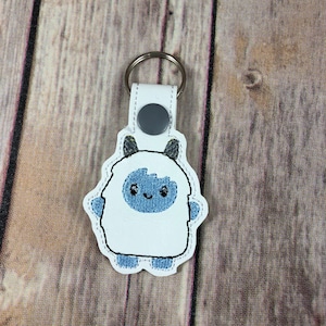 May include: White felt keychain with an embroidered blue yeti design. The yeti has a white body, blue face, and yellow horns. The keychain has a silver metal ring.
