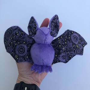 May include: A plush purple bat with black and gold patterned wings. The bat's wings feature a celestial design with zodiac symbols and stars. The bat is made of soft, fuzzy material and has small black eyes.