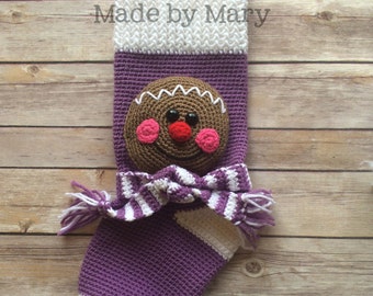 May include: Purple and white crocheted Christmas stocking with a gingerbread man face appliqué. The stocking has a white and purple striped scarf and a white tassel. The text "Made by Mary" is visible at the top of the stocking.