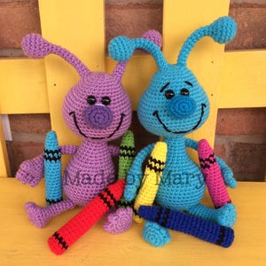 May include: Two crocheted plush toys shaped like bugs with smiling faces and large eyes. The toys are holding colorful crayons. The toys are purple and blue. "Made by Mary" is written on the bottom of the image.