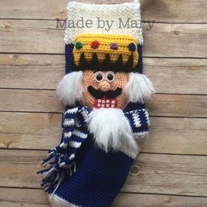 May include: A blue and white crocheted stocking with a nutcracker face, a white beard, and a yellow crown. The stocking has a white and blue striped scarf and a white tassel. The text "Made by Mary" is visible at the top of the stocking.