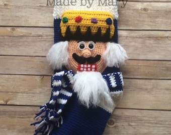 May include: A blue and white crocheted stocking with a nutcracker face, a white beard, and a yellow crown. The stocking has a white and blue striped scarf and a white tassel. The text "Made by Mary" is visible at the top of the stocking.