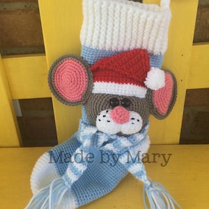 Puede incluir: Una media de Navidad de crochet con forma de ratón que lleva un gorro de Papá Noel rojo y una bufanda azul y blanca. La media es azul y blanca con un puño blanco y un pompón blanco en el gorro. El texto "Made by Mary" es visible en la media.