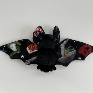 May include: A black plush bat with a patterned fabric wing. The fabric features a variety of designs including stars, snakes, books, and mushrooms.