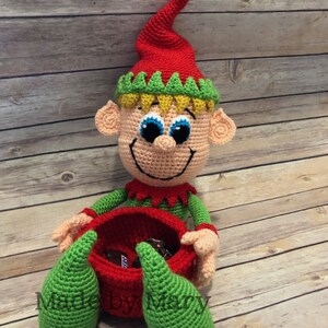 May include: A crocheted elf doll with a red hat, green and yellow trim, and a red basket. The elf is holding a green crocheted leg on each side of the basket. The basket is filled with chocolate candy bars. The text "Made by Mary" is visible on the bottom of the image.