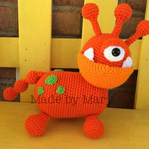 May include: A crocheted orange monster toy with green spots, one large white eye, and a toothy grin. The toy is sitting on a yellow surface.