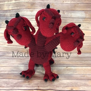 Peut inclure: Un dragon à trois têtes en crochet rouge avec des yeux et des griffes noirs. Le dragon est debout sur une surface en bois.