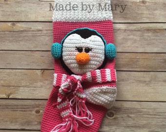 May include: A pink and white crocheted Christmas stocking with a penguin face, blue earmuffs, and a white and pink striped scarf. The stocking has the text "Made by Mary" on the top.