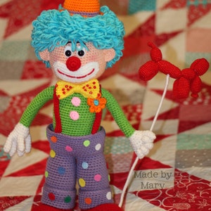 May include: A crocheted clown doll with a red nose, yellow bow tie, and colorful polka dot pants. The doll is holding a red balloon and is standing on a red, white, and blue patchwork quilt. The text "Made by Mary" is visible in the lower right corner of the image.
