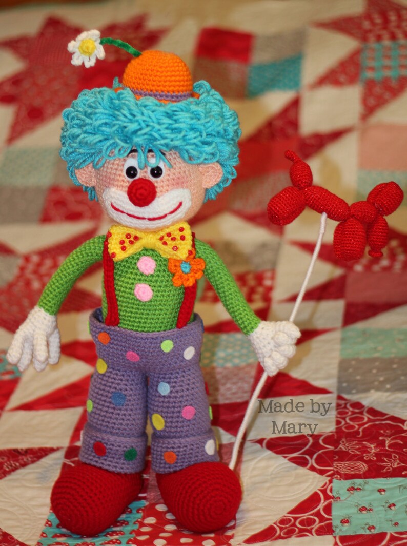 May include: A crocheted clown doll with a red nose, yellow bow tie, and colorful polka dot pants. The doll is holding a red balloon and is standing on a red, white, and blue patchwork quilt. The text "Made by Mary" is visible in the lower right corner of the image.