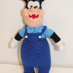 Puede incluir: Juguete de peluche de Goofy tejido a crochet, un personaje de dibujos animados de Disney. El juguete lleva un mono azul con botones blancos y una camisa azul claro. El juguete tiene una nariz negra, ojos blancos y una boca roja.