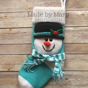 May include: A crocheted snowman stocking with a white and teal striped body, a black hat with a red holly berry, and a white and teal striped scarf. The stocking has a white loop for hanging and the text "Made by Mary" is visible on the top.