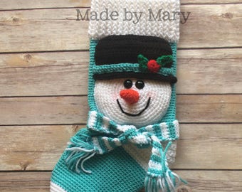 May include: A crocheted snowman stocking with a white and teal striped body, a black hat with a red holly berry, and a white and teal striped scarf. The stocking has a white loop for hanging and the text "Made by Mary" is visible on the top.