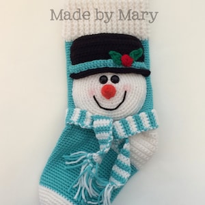 May include: A crocheted snowman stocking with a white and blue striped scarf, a black hat with a red berry and green leaves, and the text "Made by Mary".