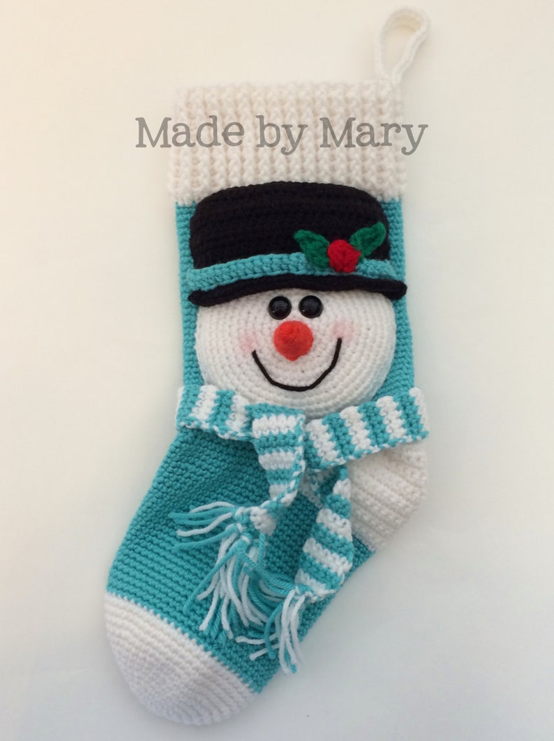 May include: A crocheted snowman stocking with a white and blue striped scarf, a black hat with a red berry and green leaves, and the text "Made by Mary".