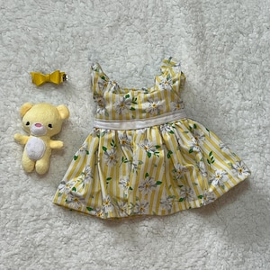 May include: A yellow and white striped dress with a floral pattern, a yellow bow hair clip, and a small yellow teddy bear. The dress has a white waistband and a ruffled neckline. The teddy bear has a pink nose and is wearing a yellow bow.