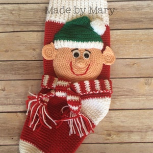 May include: Red and white crocheted Christmas stocking with a green elf face and a white pom-pom hat. The stocking has a white and red striped scarf with tassels. The stocking is labeled "Made by Mary".