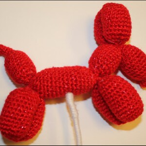 May include: A red crocheted balloon animal shaped like a dog. The dog is standing on a white stick.
