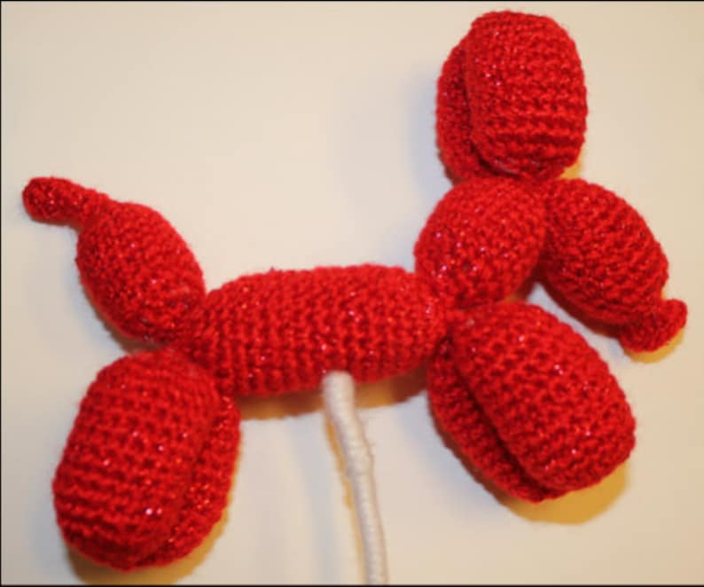 May include: A red crocheted balloon animal shaped like a dog. The dog is standing on a white stick.
