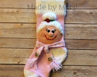 May include: Pink crocheted Christmas stocking with a white crocheted face and a white crocheted bun. The stocking has a white crocheted cuff and a white crocheted trim. The stocking has a brown button and a pink tassel. The stocking has the text "Made by Mary" on the cuff.