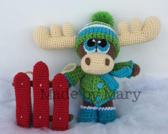 Puede incluir: Un alce de ganchillo con un atuendo de invierno verde y azul, que incluye un gorro, una bufanda y botas. El alce tiene ojos grandes y expresivos y sostiene una pequeña valla roja. El texto "Made by Mary" es visible.