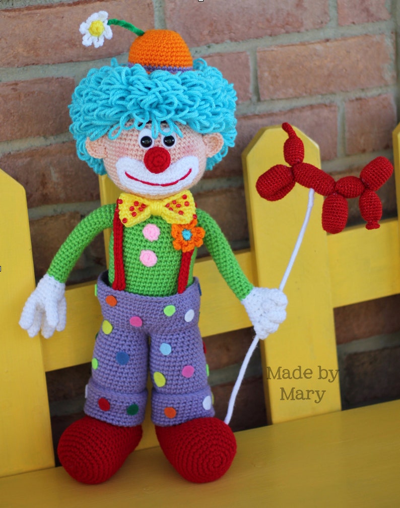 May include: A crocheted clown doll with blue hair, a red nose, and a green and purple outfit. The clown is holding a red balloon animal and is sitting on a yellow bench. Made by Mary.