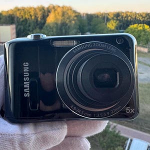 Può includere: Una fotocamera digitale Samsung nera con un prominente obiettivo Samsung Zoom Lens con zoom ottico 5x. Il barilotto dell'obiettivo riporta le specifiche 4.9-24.5 mm 1:3.5-5.9 27mm. Il corpo della fotocamera ha una finitura lucida con il logo Samsung.